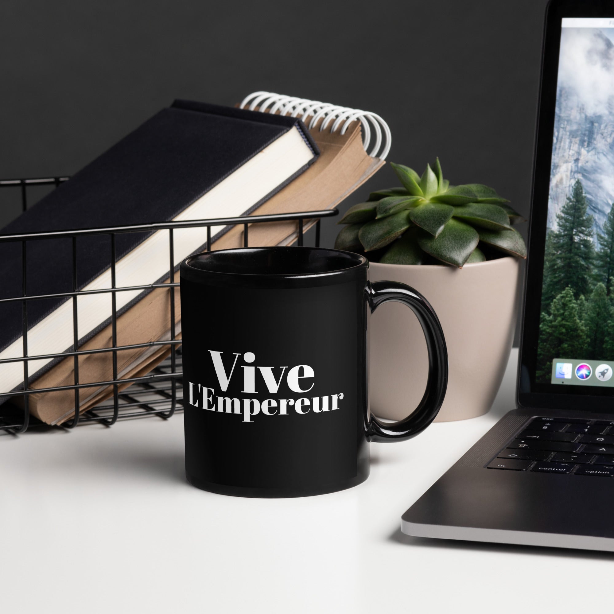 Black mug with 'Vive L'Empereur' text on a desk with laptop, books, and plant.