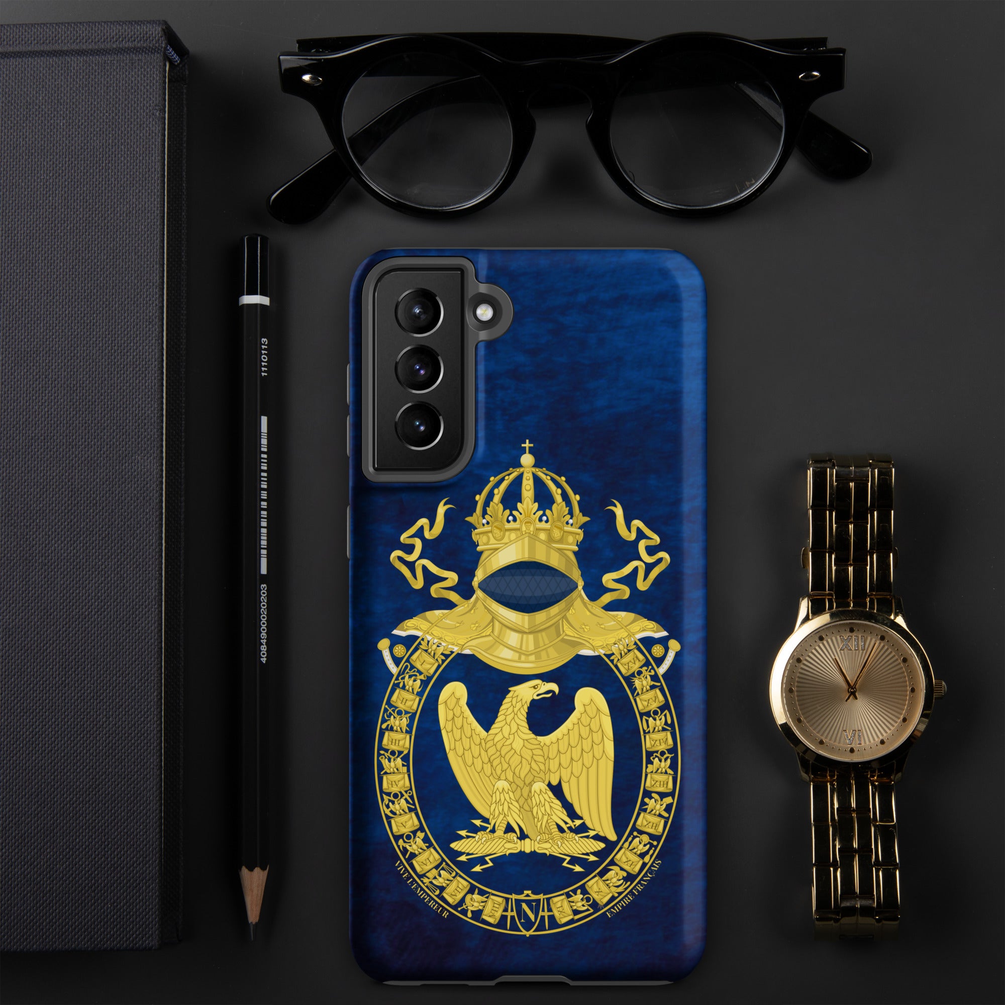 Phone case with a gold emblem on a blue background, surrounded by glasses, pen, pencil, and watch on a dark surface.